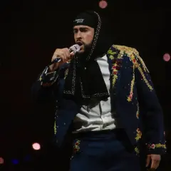 Bad Bunny w Polsce, w ozdobnej marynarce i czapce z napisem "Puerto Rico", śpiewa do mikrofonu na tle rozmytych świateł.