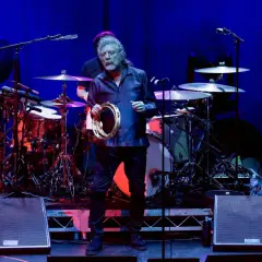 Robert Plant z zespołu zeppy zep, trzymający tamburyn na scenie, w tle perkusja i wzmacniacze.