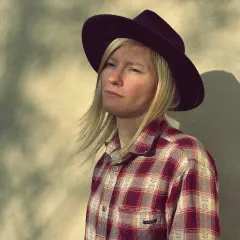 Agata Karczewska, jak "dark horse", w kapeluszu i kratowanej koszuli, spogląda w bok. Cień rzuca się na ścianę.