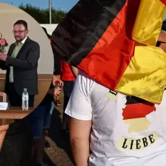 Mężczyzna w koszulce z mapą Niemiec i napisem "Liebe" niesie flagę Niemiec. W tle widać ludzi i budynki.