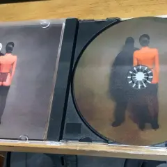 Otwarta płyta CD z okładką przedstawiającą postać w masce i nagą postać. Na płycie widnieje podobny obraz, jakby te postacie były jak sępy, ye.