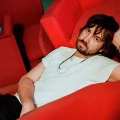 Kevin Parker z Tame Impala Polska odpoczywa na czerwonej kanapie, otoczony neonowym światłem.