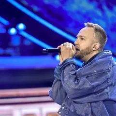 Ten typ mes wywiad na scenie, śpiewa do mikrofonu w dżinsowej kurtce, na tle niebieskich świateł.