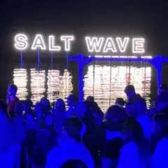 Nocny tłum na festiwalu Salt Wave w Jastarni. Neonowy napis odbija się w wodzie.