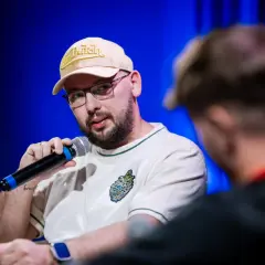 Raper Edzio w bejsbolówce Von Dutch i okularach, trzymający mikrofon, podczas panelu dyskusyjnego.