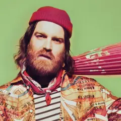 Chet Faker w czerwonej czapce i kolorowej kurtce trzyma różowy parasol.