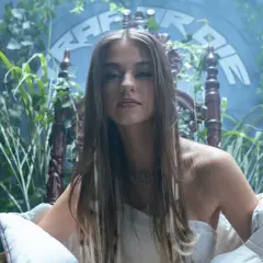 Młoda kobieta w białej sukni, otoczona bujną zielenią, siedzi na ozdobnym tronie. W tle widnieje napis "Bambi Rap".