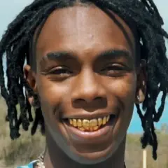 YNW Melly z dredami i złotymi zębami uśmiecha się na tle krajobrazu.