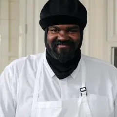 Gregory Porter w białej koszuli i czarnej czapce, z brodą, uśmiecha się w kuchni.