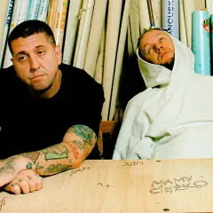 Dwóch mężczyzn, jeden z tatuażami, drugi w bluzie z kapturem, siedzi przy stole z napisem "Mamy Ciężko" i "Solar SBM".