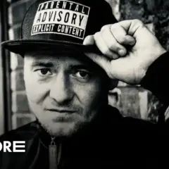 Mężczyzna w czapce z napisem "Parental Advisory Explicit Content" poprawia ją ręką. Jego spojrzenie jest intensywne, jakby mówił: "joka kaliber".