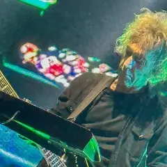 Perry Bamonte z burzą blond włosów, grający na gitarze i syntezatorze podczas koncertu, otoczony kolorowymi światłami.