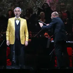 Andrea Bocelli w złotej marynarce śpiewa na koncercie w Warszawie, dyrygent prowadzi orkiestrę.
