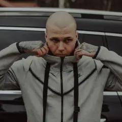 Szpaku golem, z tatuażami na rękach, poprawia kołnierz szarej bluzy Nike, opierając się o czarny samochód.