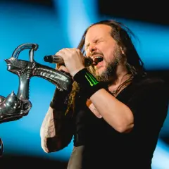 Jonathan Davis z **Korn Polska** śpiewa z pasją do mikrofonu, trzymając go na tle niebieskiego światła.
