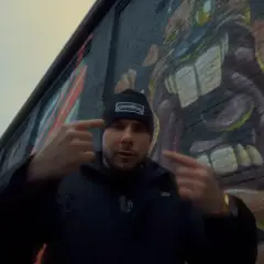 Mężczyzna w czapce z napisem "GermanStreets" wskazuje palcami na siebie, stojąc przed ścianą z graffiti. Może to być zapowiedź bonusa rpk.