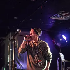 MC na scenie, w czapce i bluzie, z mikrofonem w dłoni, w stylu rap.