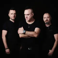 Trzech mężczyzn w czarnych ubraniach, w tym Szymon Wydra, pozuje na ciemnym tle.