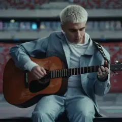 Artysta z blond włosami gra na gitarze w pustym stadionie. Zapowiada swój nowy album.