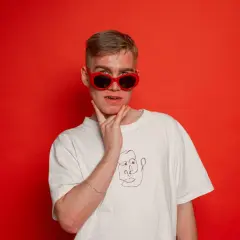 Młody mężczyzna w czerwonych okularach i białej koszulce z rysunkiem twarzy, jakby szykował się do występu koza rap.