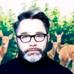 Mężczyzna w okularach z niebieskimi oczami, na tle lasu z jeleniami. Wygląda jak robag wruhme, który zgubił się w lesie.