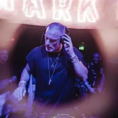 Chriss Jaxx w akcji, miksuje muzykę w klubie. W tle neonowy napis i ludzie bawiący się w rytm jego setu.