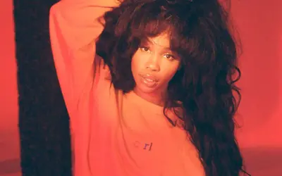 SZA w pomarańczowej bluzie z napisem "crl", z ręką we włosach na tle czerwonym.