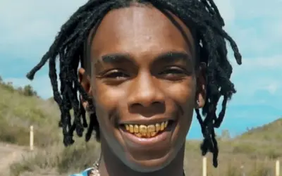 YNW Melly z dredami i złotymi zębami uśmiecha się na tle krajobrazu.