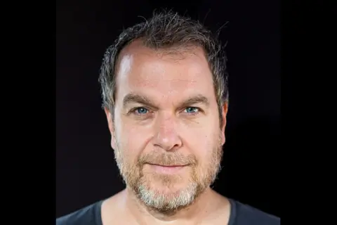 Marcel Woods, z niebieskimi oczami i siwiejącą brodą, patrzy prosto w obiektyw.
