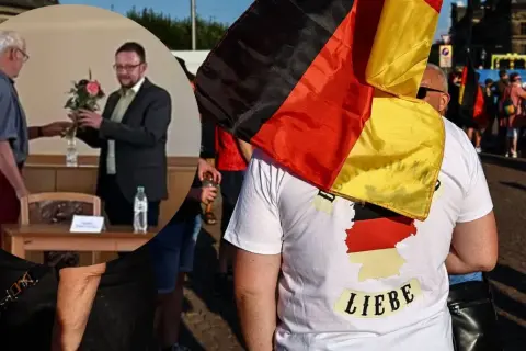 Mężczyzna w koszulce z mapą Niemiec i napisem "Liebe" niesie flagę Niemiec. W tle widać ludzi i budynki.
