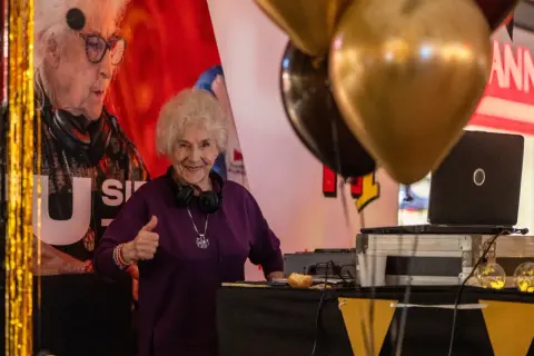 Uśmiechnięta starsza pani z słuchawkami na szyi, dająca kciuk w górę przy stanowisku DJ-a. To DJ Rush w akcji!