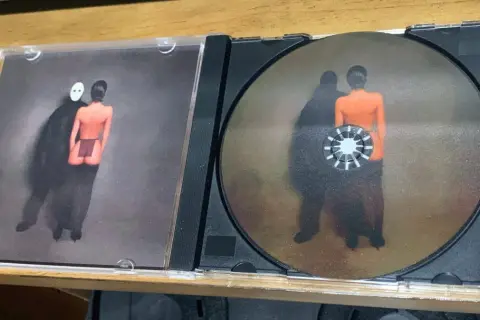 Otwarta płyta CD z okładką przedstawiającą postać w masce i nagą postać. Na płycie widnieje podobny obraz, jakby te postacie były jak sępy, ye.