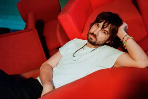 Kevin Parker z Tame Impala Polska odpoczywa na czerwonej kanapie, otoczony neonowym światłem.