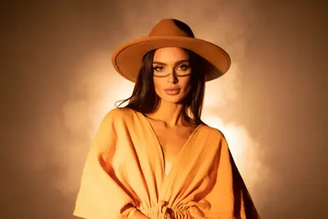 DJ Korolova w kapeluszu i pomarańczowej sukience, z ozdobnym makijażem na twarzy, emanuje tajemniczością i stylem.
