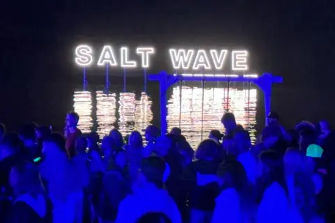 Nocny tłum na festiwalu Salt Wave w Jastarni. Neonowy napis odbija się w wodzie.