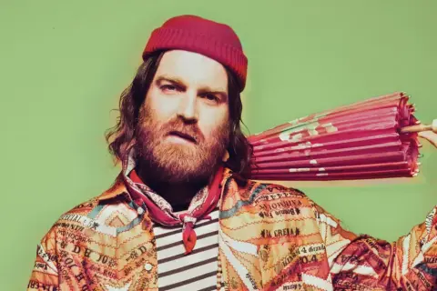 Chet Faker w czerwonej czapce i kolorowej kurtce trzyma różowy parasol.
