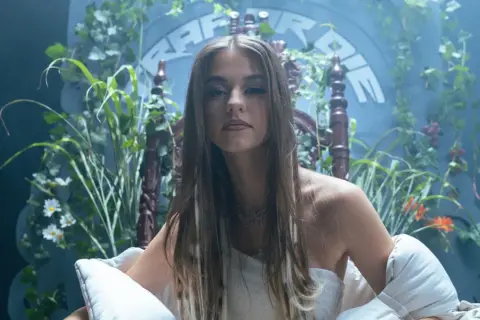 Młoda kobieta w białej sukni, otoczona bujną zielenią, siedzi na ozdobnym tronie. W tle widnieje napis "Bambi Rap".