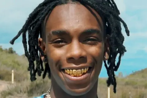 YNW Melly z dredami i złotymi zębami uśmiecha się na tle krajobrazu.