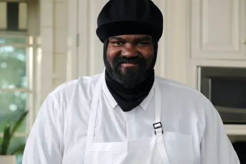 Gregory Porter w białej koszuli i czarnej czapce, z brodą, uśmiecha się w kuchni.