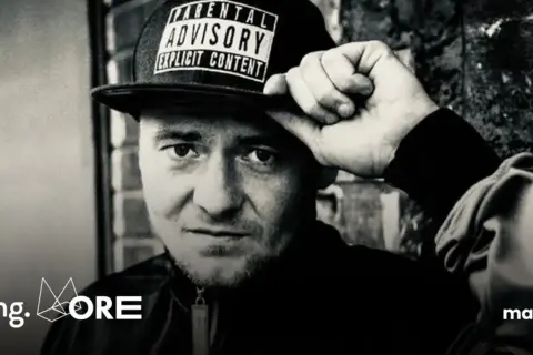 Mężczyzna w czapce z napisem "Parental Advisory Explicit Content" poprawia ją ręką. Jego spojrzenie jest intensywne, jakby mówił: "joka kaliber".