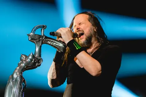 Jonathan Davis z **Korn Polska** śpiewa z pasją do mikrofonu, trzymając go na tle niebieskiego światła.