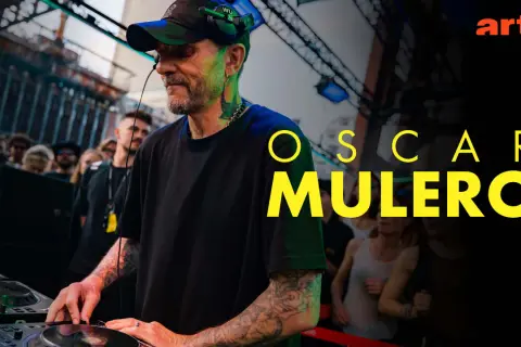 Oscar Mulero, DJ z tatuażami, miksuje muzykę na żywo, podczas gdy tłum ludzi bawi się w tle. Arte.