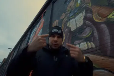 Mężczyzna w czapce z napisem "GermanStreets" wskazuje palcami na siebie, stojąc przed ścianą z graffiti. Może to być zapowiedź bonusa rpk.