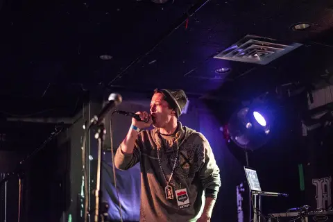 MC na scenie, w czapce i bluzie, z mikrofonem w dłoni, w stylu rap.