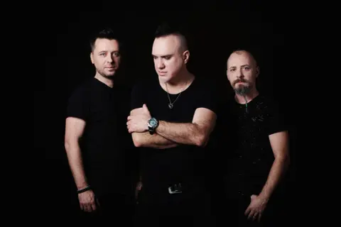 Trzech mężczyzn w czarnych ubraniach, w tym Szymon Wydra, pozuje na ciemnym tle.