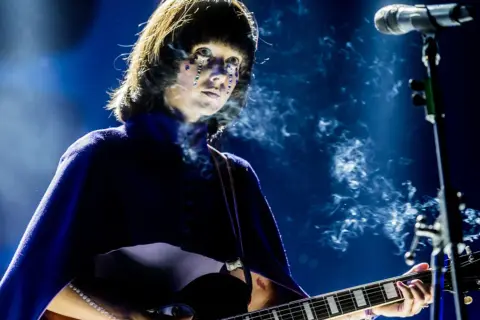 Artystka z błyszczącymi łzami na twarzy gra na gitarze podczas MTV Unplugged Polska. Dym unosi się wokół niej.
