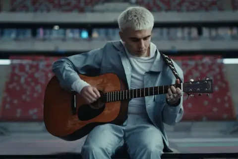 Artysta z blond włosami gra na gitarze w pustym stadionie. Zapowiada swój nowy album.
