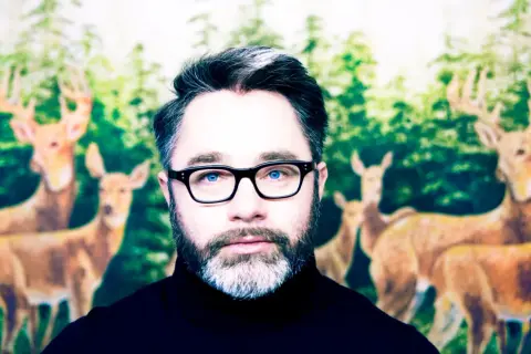 Mężczyzna w okularach z niebieskimi oczami, na tle lasu z jeleniami. Wygląda jak robag wruhme, który zgubił się w lesie.