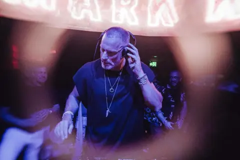 Chriss Jaxx w akcji, miksuje muzykę w klubie. W tle neonowy napis i ludzie bawiący się w rytm jego setu.