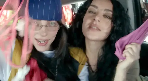 Dwie młode kobiety, jedna z nich przypomina brata Billie Eilish, trzymają w rękach kolorowe tkaniny, bawiąc się w samochodzie.